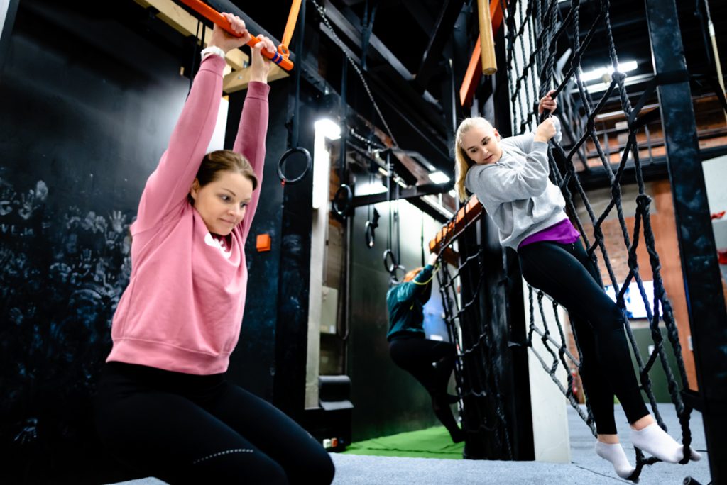 Kaksi ryhmän jäsentä suorittavat tehtäväradan tehtävää köysiseinässä ja tangossa roikkuen Wasa Up kiipeilysalissa