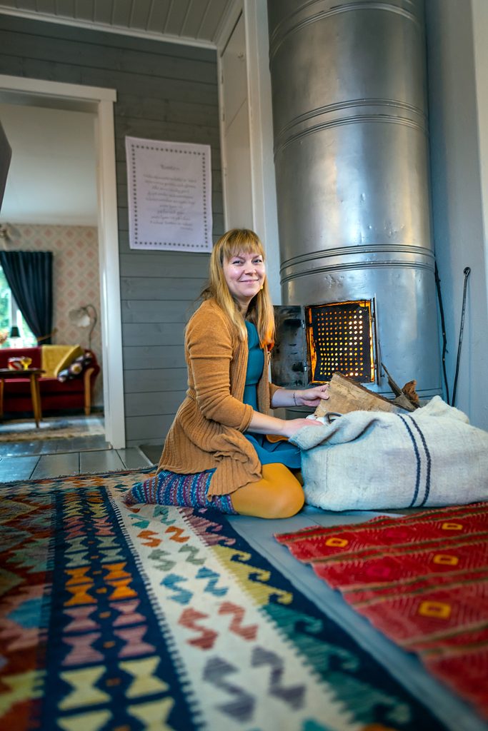 Johanna istuu lattialla ruokailuhuoneessa ja lisää puita kakluuniuunin pesään